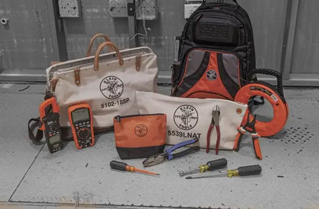 klein testers, screwdrivers, plies, and bags arranged on display on the ground